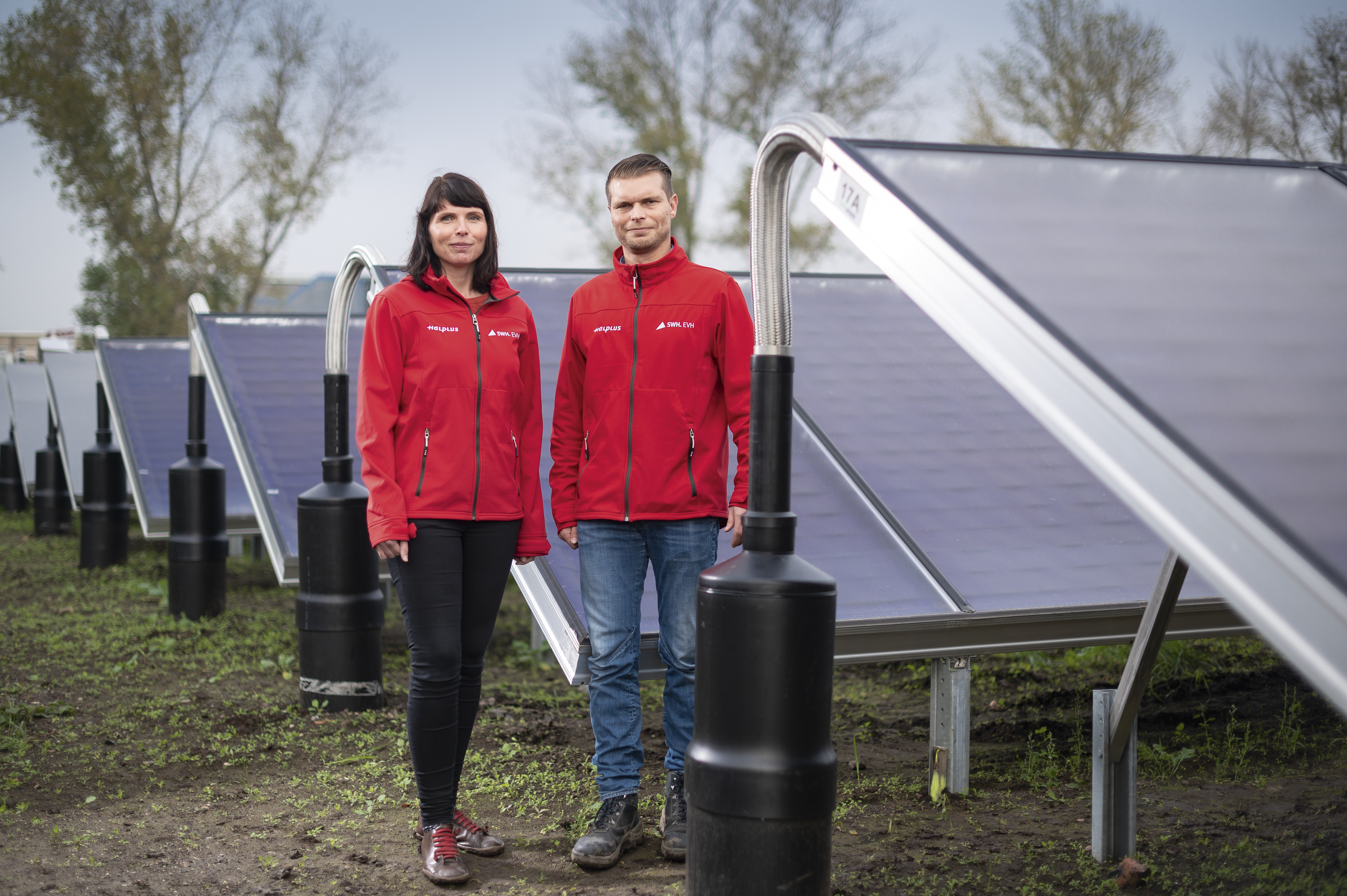 Auf der rechten Seite stehen große Solarmodule, links davon zwei Mitarbeitende der EVH, eine Frau und ein Mann, die jeweils eine leuchtend rote Jacke tragen.