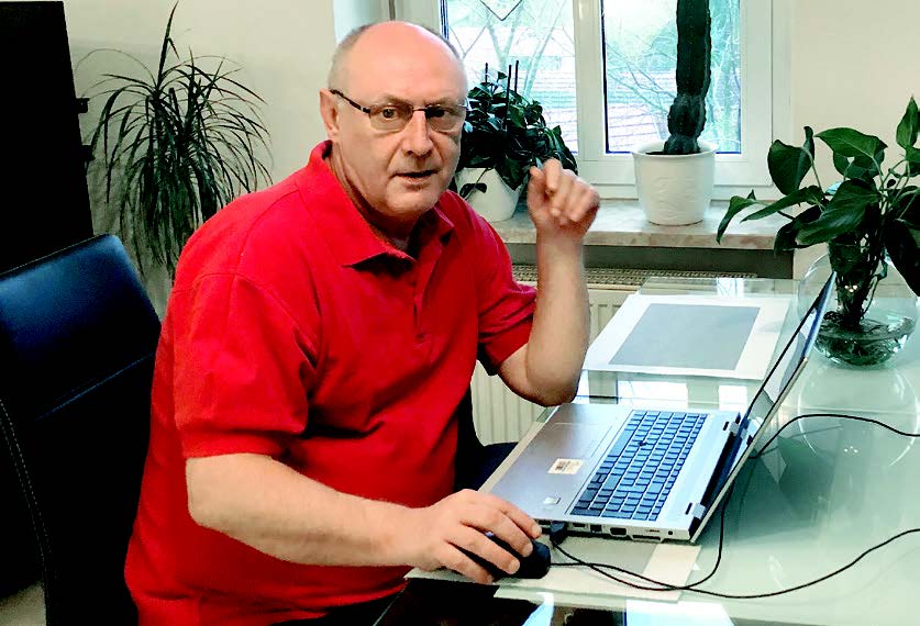 In der linken Bildhälfte sitzt ein Mann in einem roten Polohemd und mit Brille an seinem Schreibtisch. Er schaut in die Kamera, während er mit seiner rechten Hand die Maus seines Laptops bedient. 