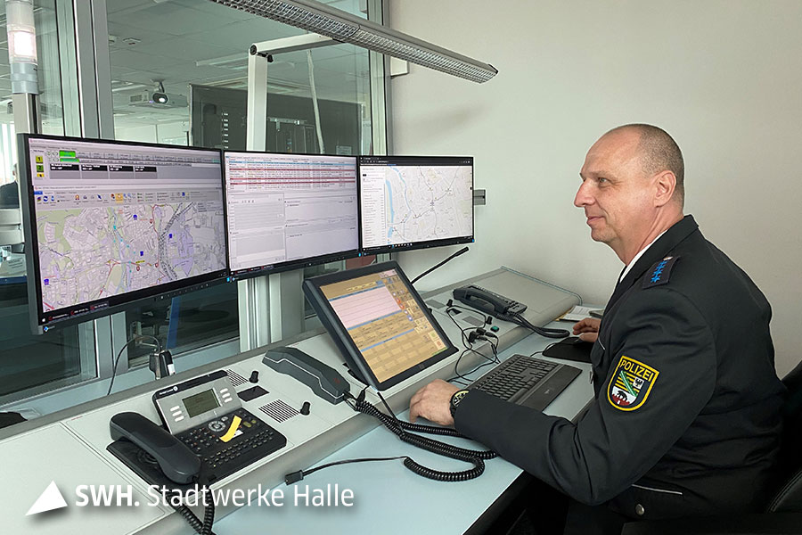 Ein Mann mit Polizeiuniform sitzt an einem Schreibtisch und schaut vor sich auf drei Monitore mit Straßenkarten. 