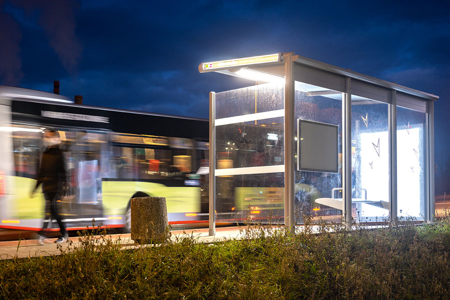 Ein Bus fährt von der umbenannten Haltestelle ab. Es ist Abend und der Himmel dunkelblau. Eine dunkel gekleidete Person bewegt sich an der Haltestelle. Die Haltestelle ist beleuchtet.