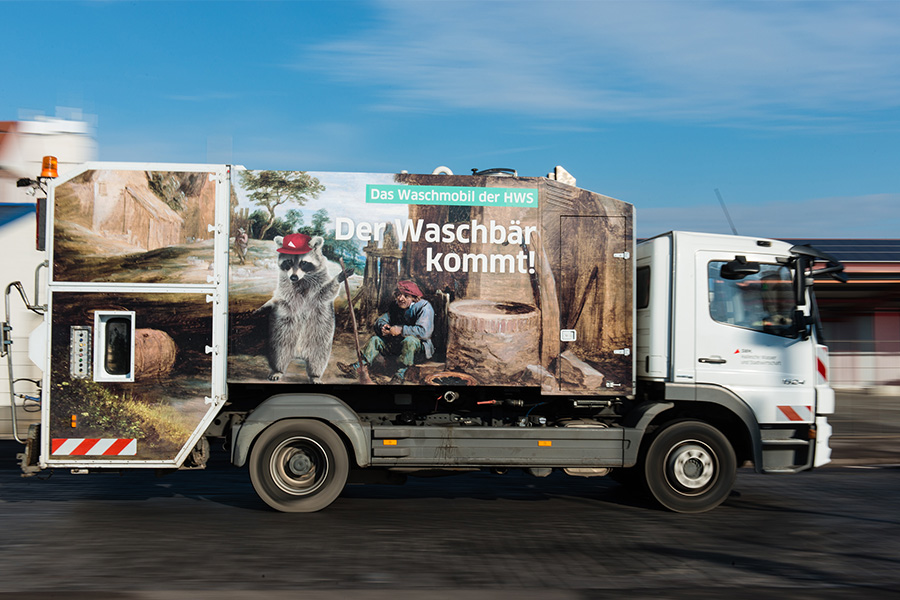 Der Waschbär - ein Auto der HWS, welches Mülltonnen reinigt fährt über die Straße. Der Hintergrund ist unscharf.