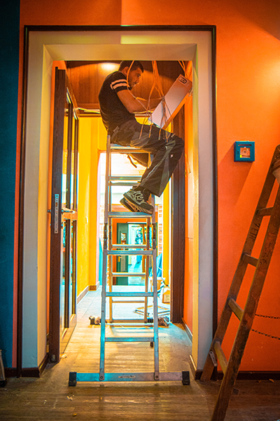 Das Foto zeigt einen Türrahmen und dahinter einen Mann, der auf einer Leiter sitzt und an Kabeln arbeitet.