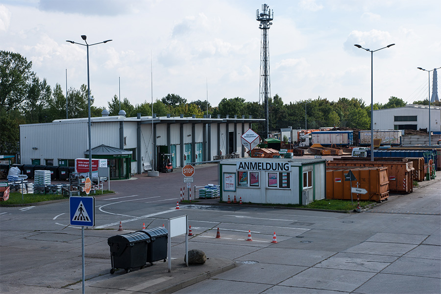 Ein Bild der des Annahmecontainers des Wertstoffmarktes aus einer leichten Vogelperspektive. Der Container ist weiß und hat die Überschrift "Abmeldung". Daneben steht ein Stopp-Schild, an dem Die Autos halten müssen. Im Hintergrund ist eine Art Lagerhaus zu sehen.