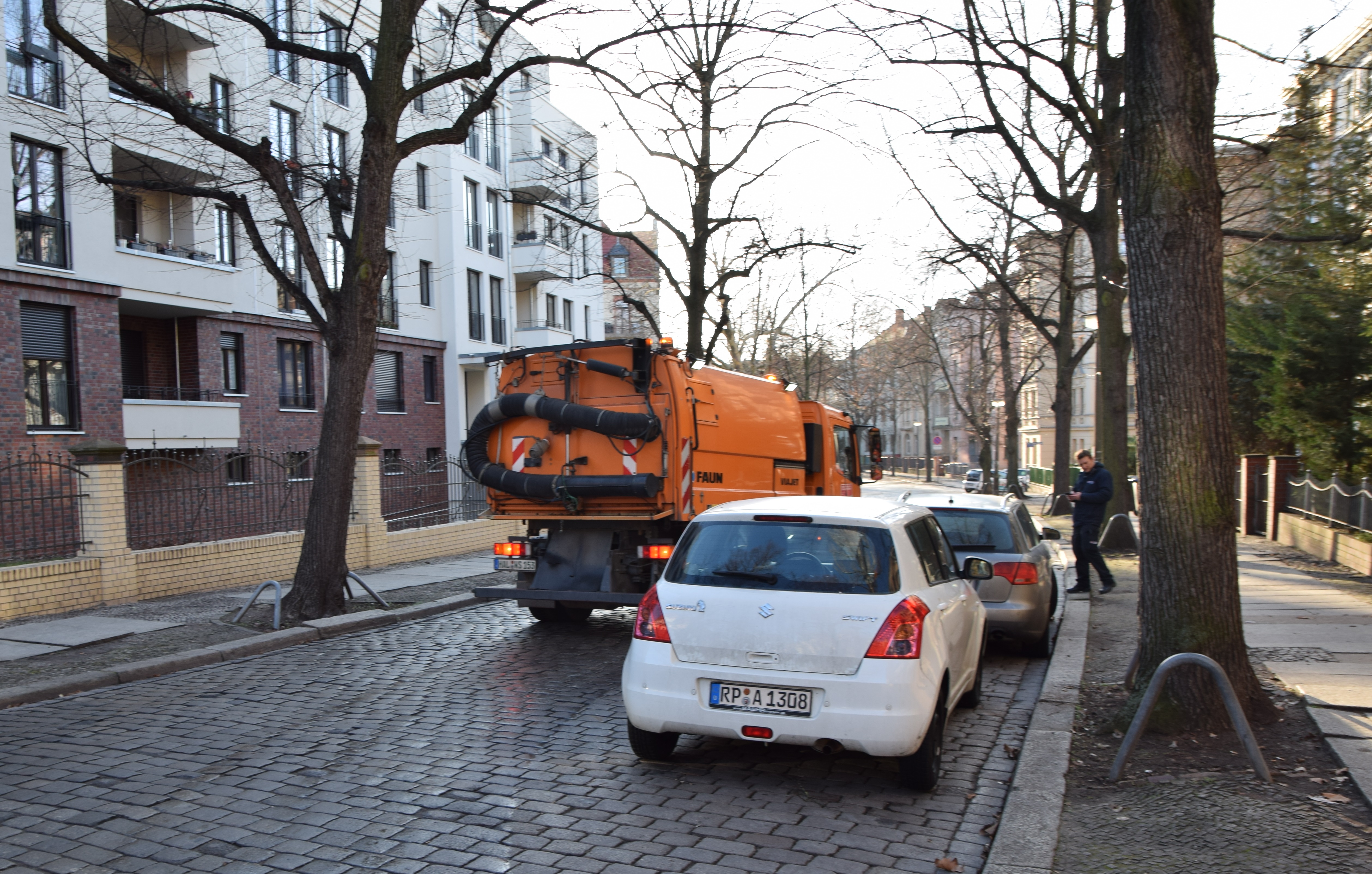 Auf dem Bild ist eine gepflasterte Straße in einem Wohngebiet abgebildet. Am rechten Fahrbahnrand parken ein weißes und ein silbernes Auto, die orangefarbene Straßenreinigungsmaschine der HWS muss am linken Fahrbahnrand fahren, um den Autos auszuweichen.