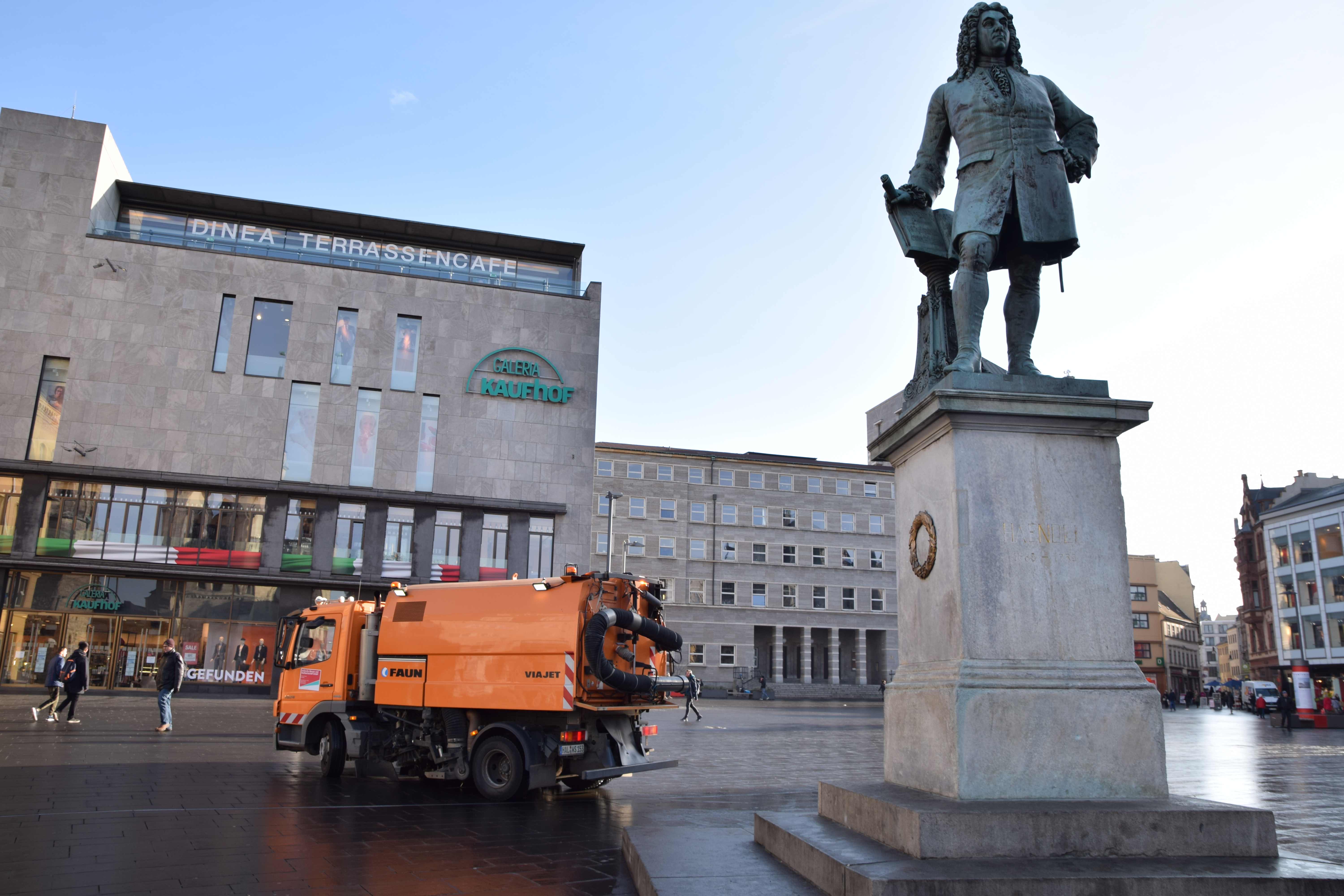 Hier ist eine Straßenreinigungsmaschine der HWS auf dem Halleschen Marktplatz abgebildet. Im Hintergrund befindet sich ein großes Einkaufszentrum, im Vordergrund das Händel-Denkmal.