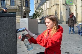 Eine Dame geht vor dem Brunnen in die Knie. Sie hält eine Flasche an die Flaschenfüll-Armatur und befüllt sie mit Trinkwasser.