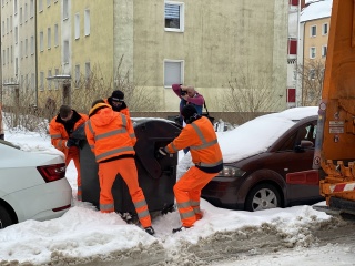 In der Mitte des Bildes befinden sich vier Männer, die in orangefarbenen Anzügen und mit Mützen und Mundschutz gekleidet sind und einen grauen Container durch Schneeberge zwischen zwei Autos hindurch ziehen und schieben.