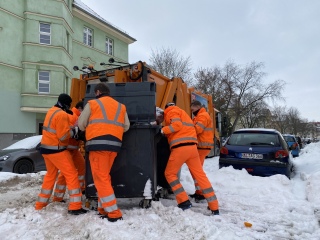 Dieses Bild zeigt mittig fünf Männer in orangefarbenen Anzügen, die einen grauen Container über eine schneebedeckte Straße hin zu einem orangefarbenen Fahrzeug der Müllabfuhr schieben.