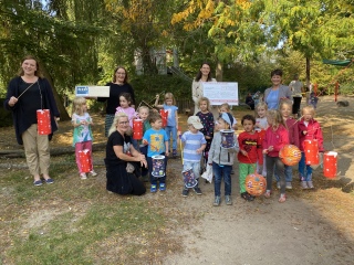 Das Bild wurde in einem Garten mit Spielgeräten und Bäumen aufgenommen und zeigt im Hintergrund vier Frauen und im Vordergrund 15 Kindergartenkinder, die viele bunte Laternen in den Händen halten.
