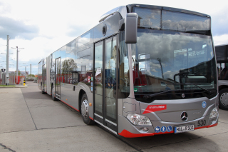 Ein silberfarbener Bus trägt über dem rechten Scheinwerfer eine rote Beklebung. Auf ihr steht der Name Anselma Heine. Der Himmel ist wolkenbehangen.