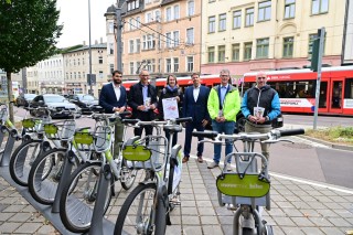 Das Bild zeigt eine Gruppe von sechs Personen, die neben einer Reihe von Mietfahrrädern von movemix stehen. Einige der Personen halten Broschüren in den Händen. Im Hintergrund sind Straßenbahnschienen auf welchen eine rote Straßenbahn der HAVAG fährt zusehen, sowie mehrstöckige Gebäude.