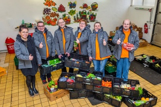 Die Helfer und Helferinnen der Tafel stehen in der Anlieferzone inmitten von Kisten mit lebensmitteln. Dabei tragen sie schon ihre neuen Softshelljacken 