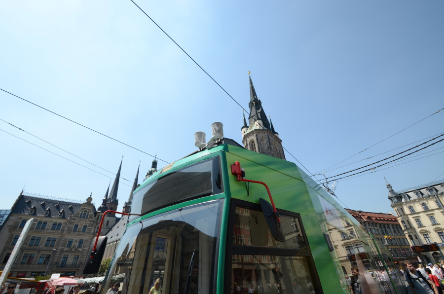 Das Bild ist von unten nach oben aufgenommen und zeigt die Fahrerkabine einer grünen Straßenbahn. Auf der Bahn befinden sich kleine graue Zylinder, die Messfühler der Bahn. Im Hintergrund ist die Marienkirche von Halle zu sehen.
