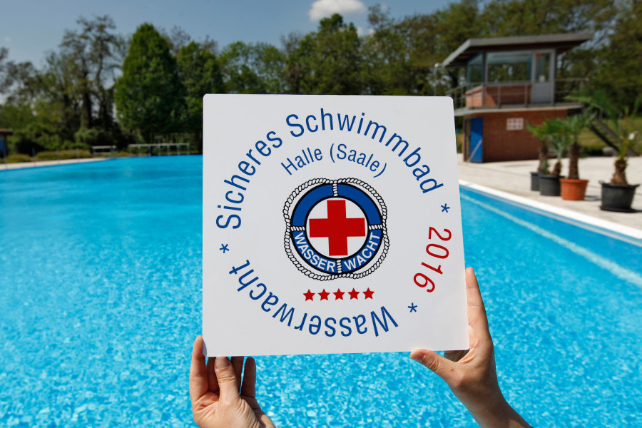 In der Mitte des Bildes befindet sich ein weißes Schild mit der Aufschrift "Sicheres Schwimmbad Halle (Saale) * 2016 * Wasserwacht". Es wird von zwei Händen gehalten. Im Hintergrund befindet sich das Schwimmbecken des Freibads Saline.