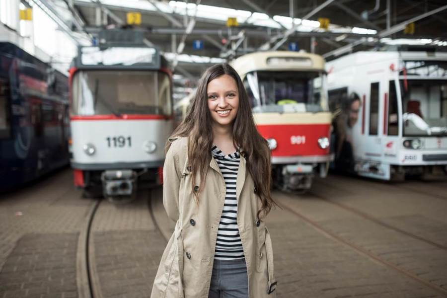 Das Bild zeigt mittig eine lächelnde junge Frau mit langen braunen Haaren, einem schwarz-weiß geringelten Oberteil und einem beigen Mantel. Sie steht im Betriebshof Rosengarten und hinter ihr befinden sich drei verschiedene Straßenbahnzüge, zwei alte Tatras und eine moderne Bahn.