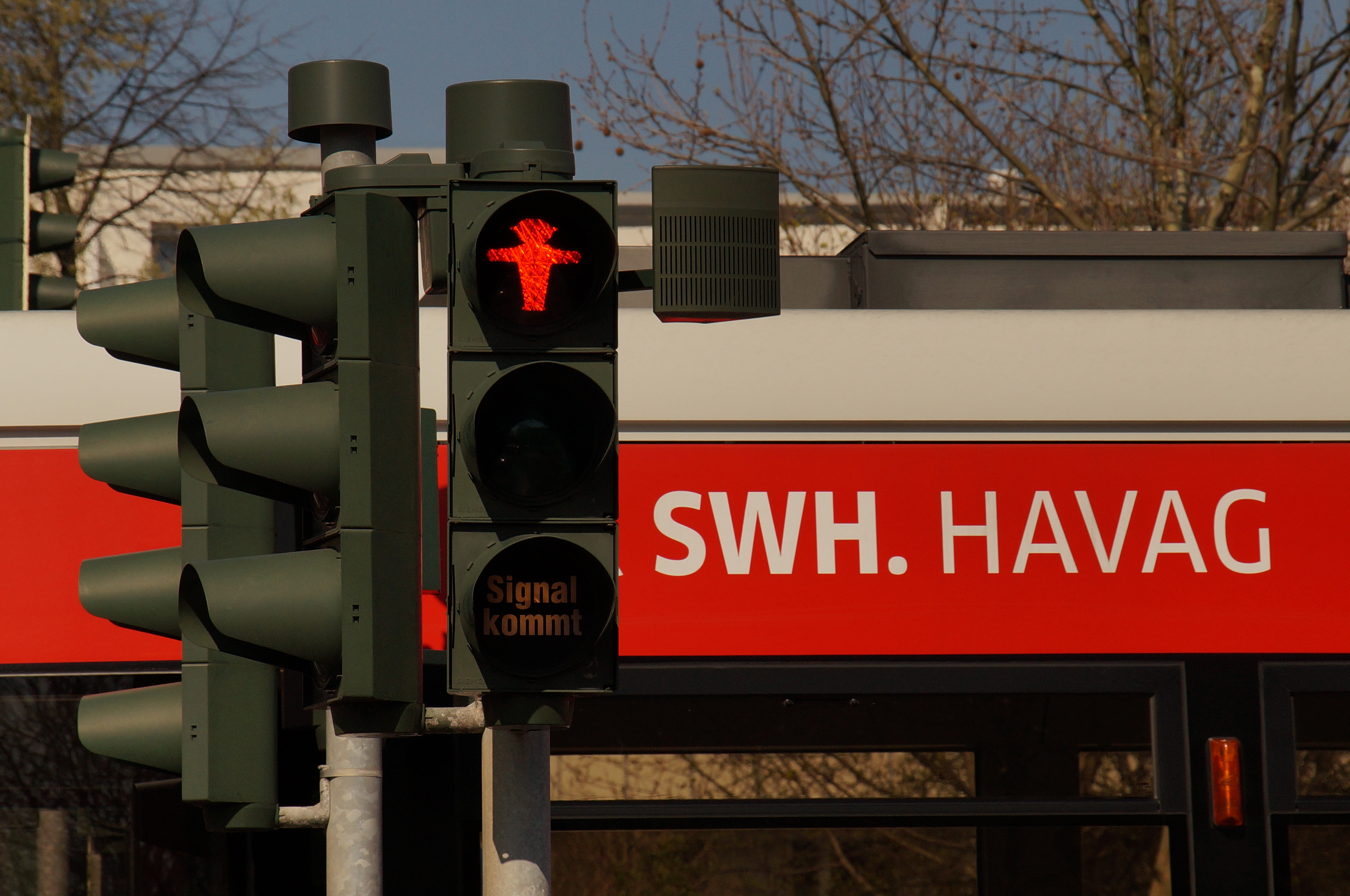 Die linke Bildhälfte zeigt eine Fußgängerampel, auf der das rote Männchen leuchtet und daneben einen Tonsignalgeber, ein kleiner grauer Kasten, der an der Ampel befestigt ist. Auf der rechten Bildhälfte befindet sich ein Bus mit der Aufschrift "SWH.HAVAG".